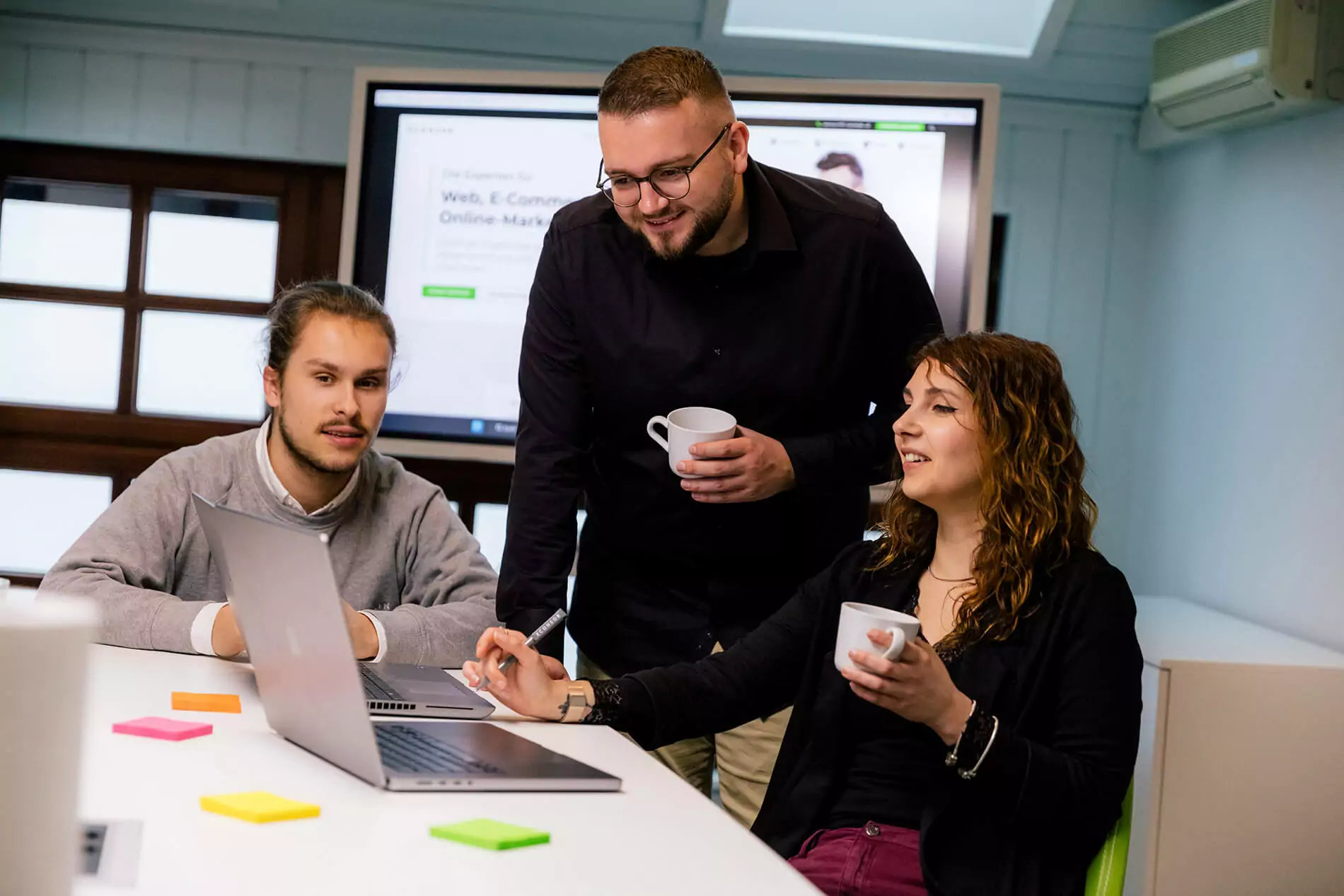Eine Gruppe von Geschäftsleuten arbeitet um einen Laptop herum, mit Haftnotizen auf dem Tisch und einem großen Bildschirm, auf dem Inhalte angezeigt werden.