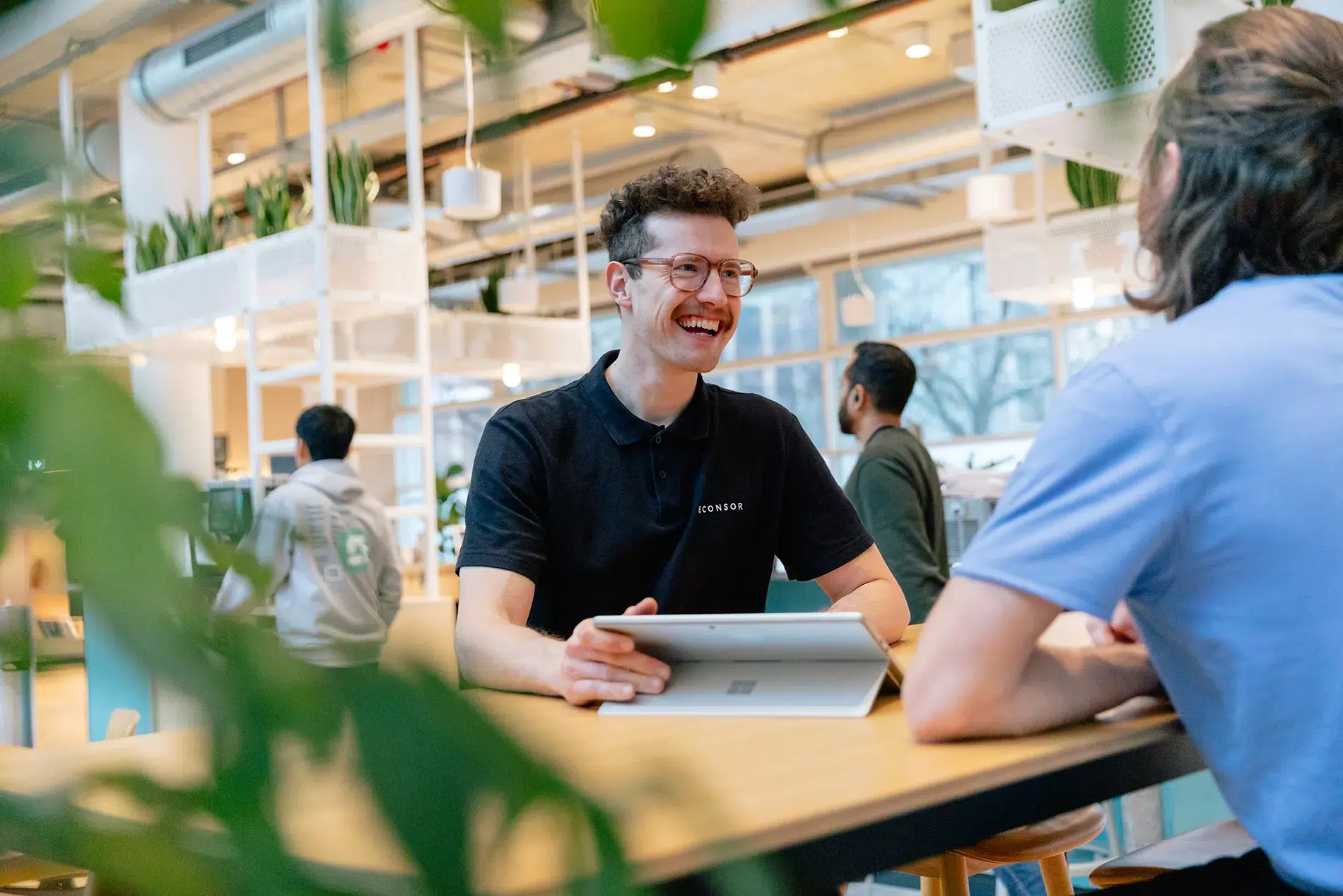 Ein Mann im schwarzen 'ECONSOR'-Poloshirt sitzt mit einem Tablet an einem Holztisch in einem modernen Büro mit viel Pflanzen und natürlichem Licht. Er lacht und unterhält sich mit einer anderen Person, während im Hintergrund weitere Mitarbeiter arbeiten.