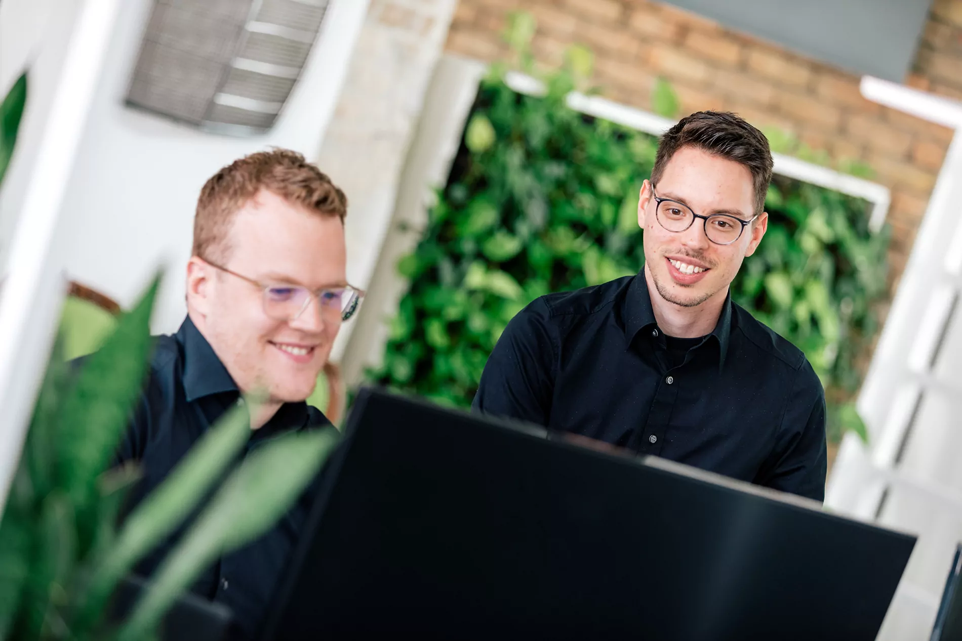 Zwei Fachleute in schwarzen Hemden arbeiten an einem Computer, umgeben von Grün in einem modernen Arbeitsraum.