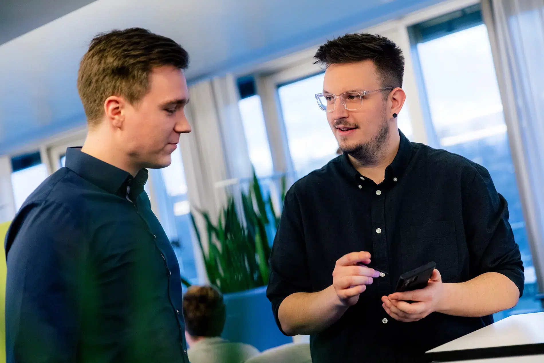 Zwei Personen führen in einem Büro ein Gespräch. Einer hält ein Telefon und gestikuliert, während der andere zuhört. Im Hintergrund sind Zimmerpflanzen und große Fenster zu sehen.