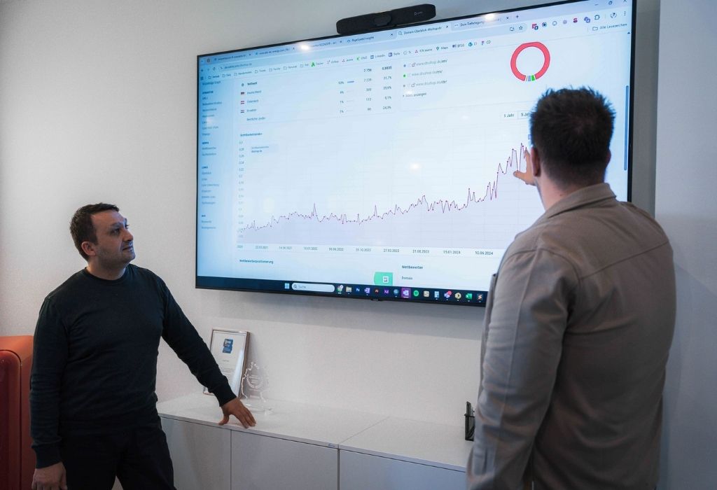 TYPO3 Besprechung TV – Zwei Mitarbeiter betrachten eine Traffic-Analyse mit steigender Kurve Zwei Mitarbeiter betrachten auf einem großen Bildschirm eine Traffic-Analyse mit steigender Kurve.