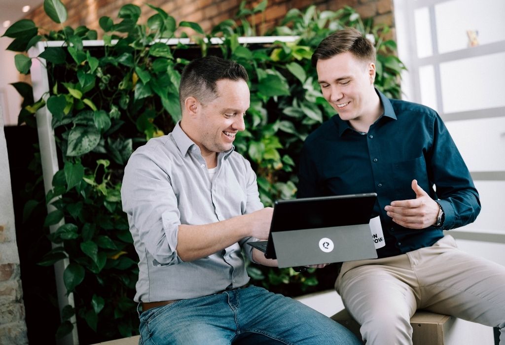 Zwei Männer sitzen entspannt vor einer begrünten Wand und besprechen etwas an einem Laptop. Beide lachen, während sie auf den Bildschirm schauen. Die Szene zeigt ein freundliches und konstruktives Arbeitsgespräch in einer modernen Agenturumgebung.
