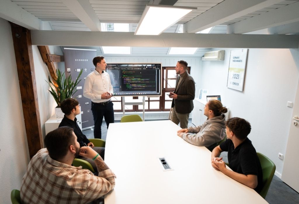 Effiziente Zusammenarbeit im TYPO3 Besprechungsraum Mehrere Personen sitzen in einem Besprechungsraum mit großem Tisch und Bildschirm. Zwei Männer präsentieren Codes auf dem Monitor, während die anderen aufmerksam zuhören. Die Szene zeigt konzentrierte Teamarbeit und technische Abstimmung.