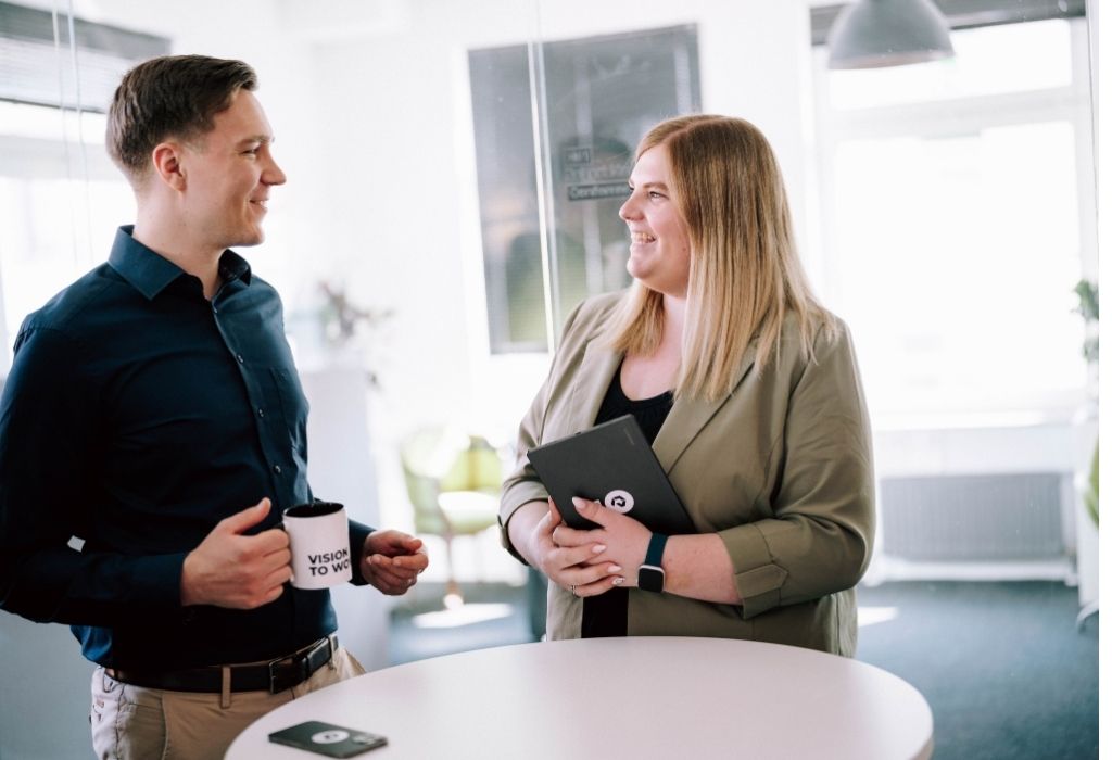 Ein Mann und eine Frau stehen an einem weißen Stehtisch, lächeln sich an und sprechen miteinander. Beide wirken entspannt und engagiert, im Hintergrund scheint helles Tageslicht durch die Fenster. Der Raum wirkt modern und offen.
