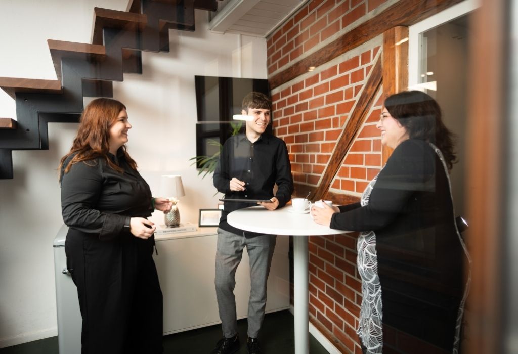 Drei Personen stehen in einem modernen Büro vor einer roten Backsteinwand und unterhalten sich lächelnd an einem hohen Stehtisch. Die Atmosphäre wirkt locker und kollegial, im Hintergrund sind eine Holztreppe und dezente Büroeinrichtung zu sehen.