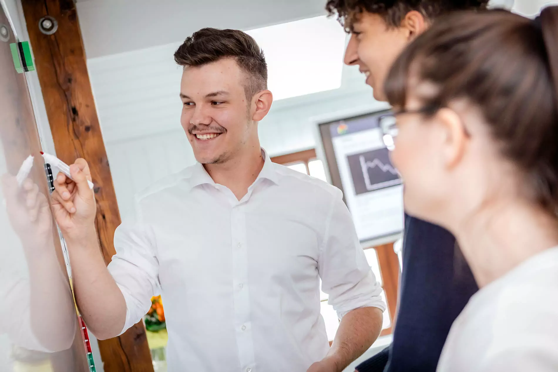 TYPO3-Agentur in Heidelberg - Fachleute in einem Meeting Eine Person in einem hellen Hemd schreibt auf einem Whiteboard, während zwei andere in einem hellen, modernen Besprechungsraum mit einem Bildschirm im Hintergrund zusehen.