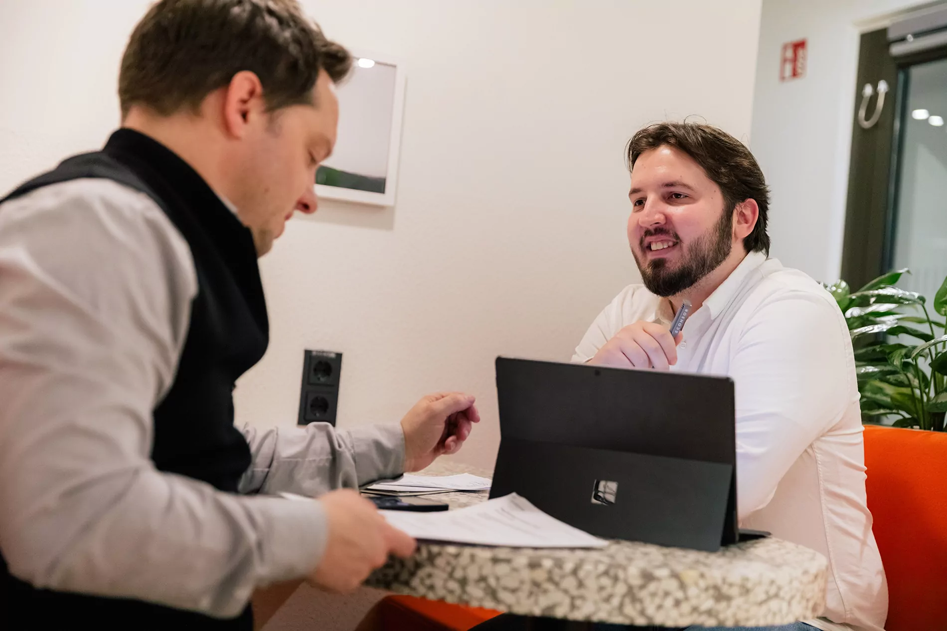Ihre TYPO3-Agentur – Expertenmeeting bei ECONSOR zur Angebots erstellung Zwei Personen sitzen an einem kleinen Tisch und besprechen Dokumente, im Hintergrund stehen ein Laptop und eine Grünpflanze.