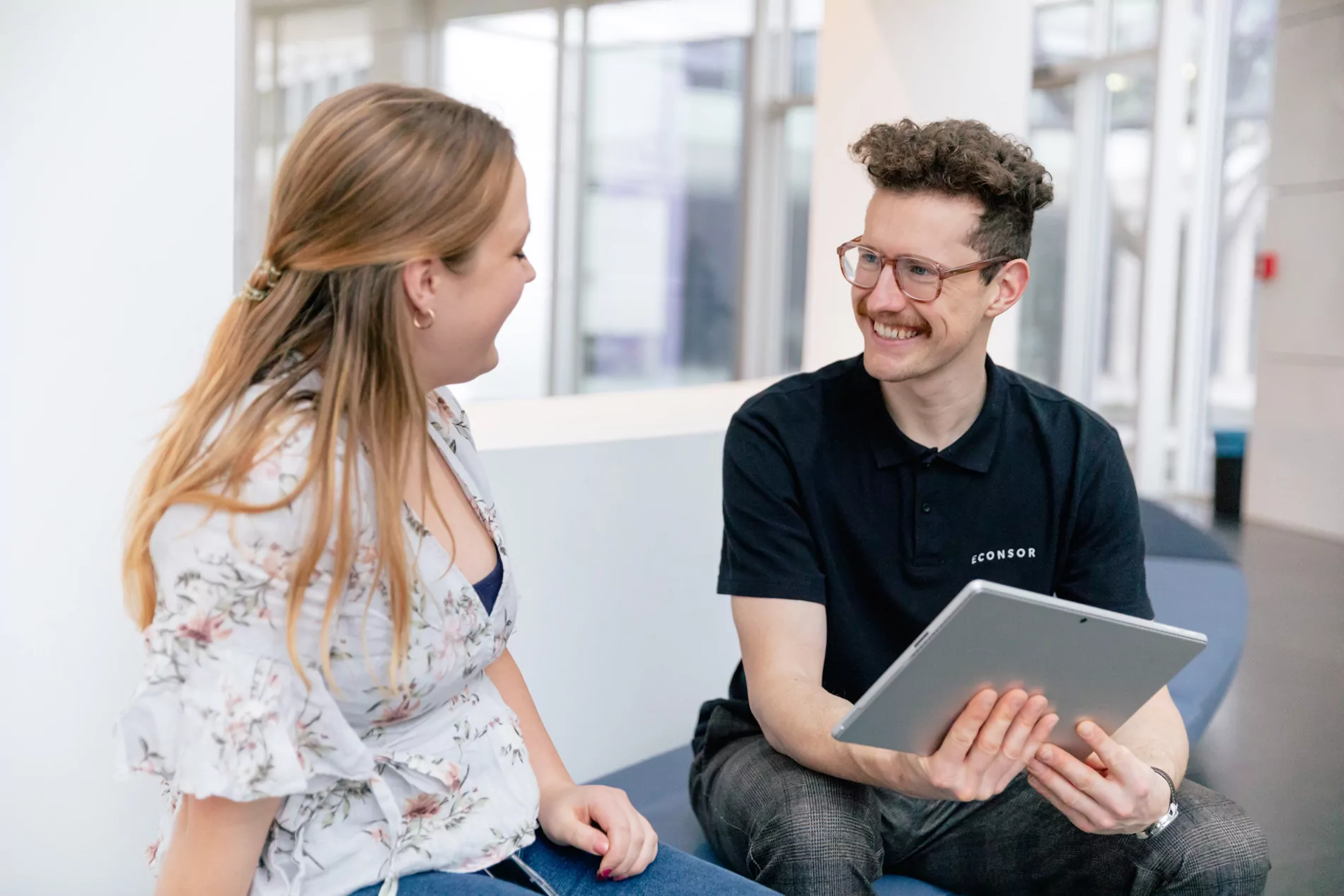 TYPO3-Agentur in München - Zwei Experten besprechen sich Ein Mann und eine Frau sitzen in einem modernen Bürogebäude und führen ein freundliches Gespräch. Der Mann hält ein Tablet in den Händen und trägt ein schwarzes Poloshirt mit der Aufschrift 'ECONSOR'.