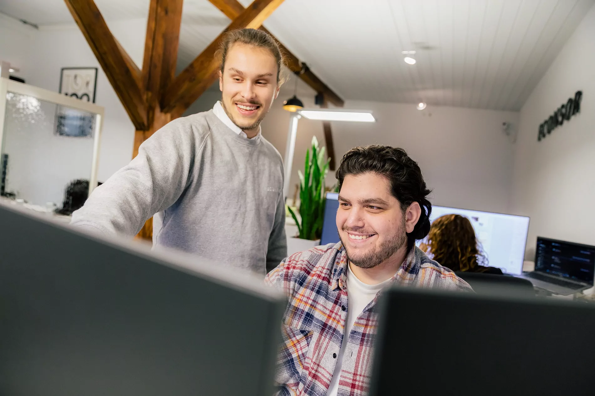 TYPO3-Agentur in Nürnberg - Zwei Kollegen im Austausch Zwei Männer schauen auf Computerbildschirme und lächeln in einem Büro mit Holzbalken und einer Pflanze im Hintergrund.