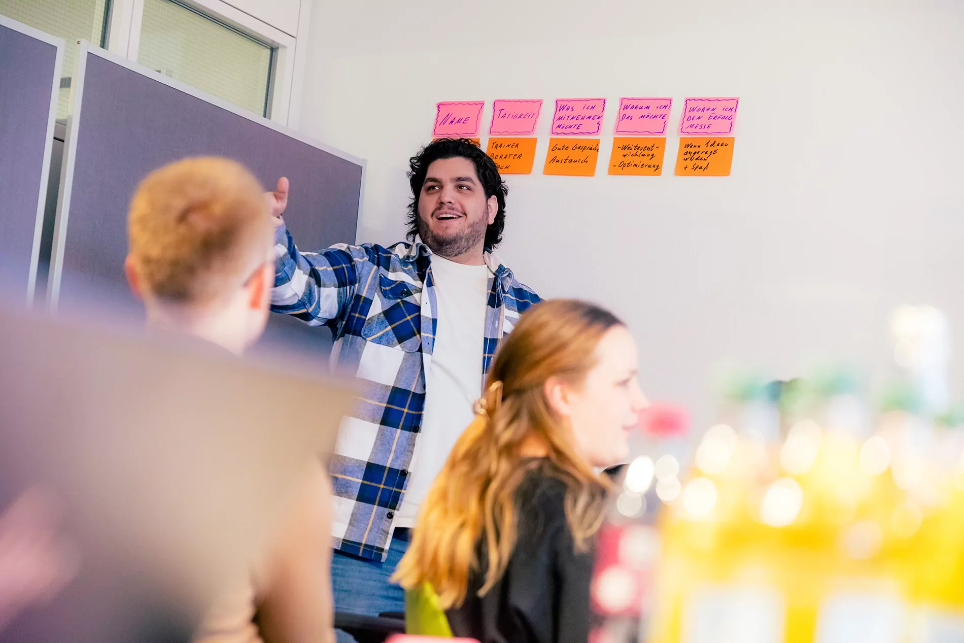 Eine Person in einem karierten Hemd gestikuliert, während sie vor einer Gruppe mit bunten Haftnotizen an der Wand präsentiert.