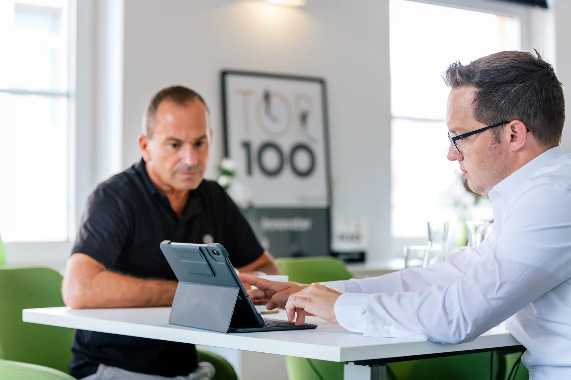 TYPO3-Agentur in Heilbronn - Experte präsentiert Ergebnis Ein Fachmann zeigt einem Kunden etwas auf einem Tablet in einer hellen, modernen Büroumgebung. Der Kunde hört aufmerksam zu. Zur Dekoration gehört ein „Top 100“-Poster.