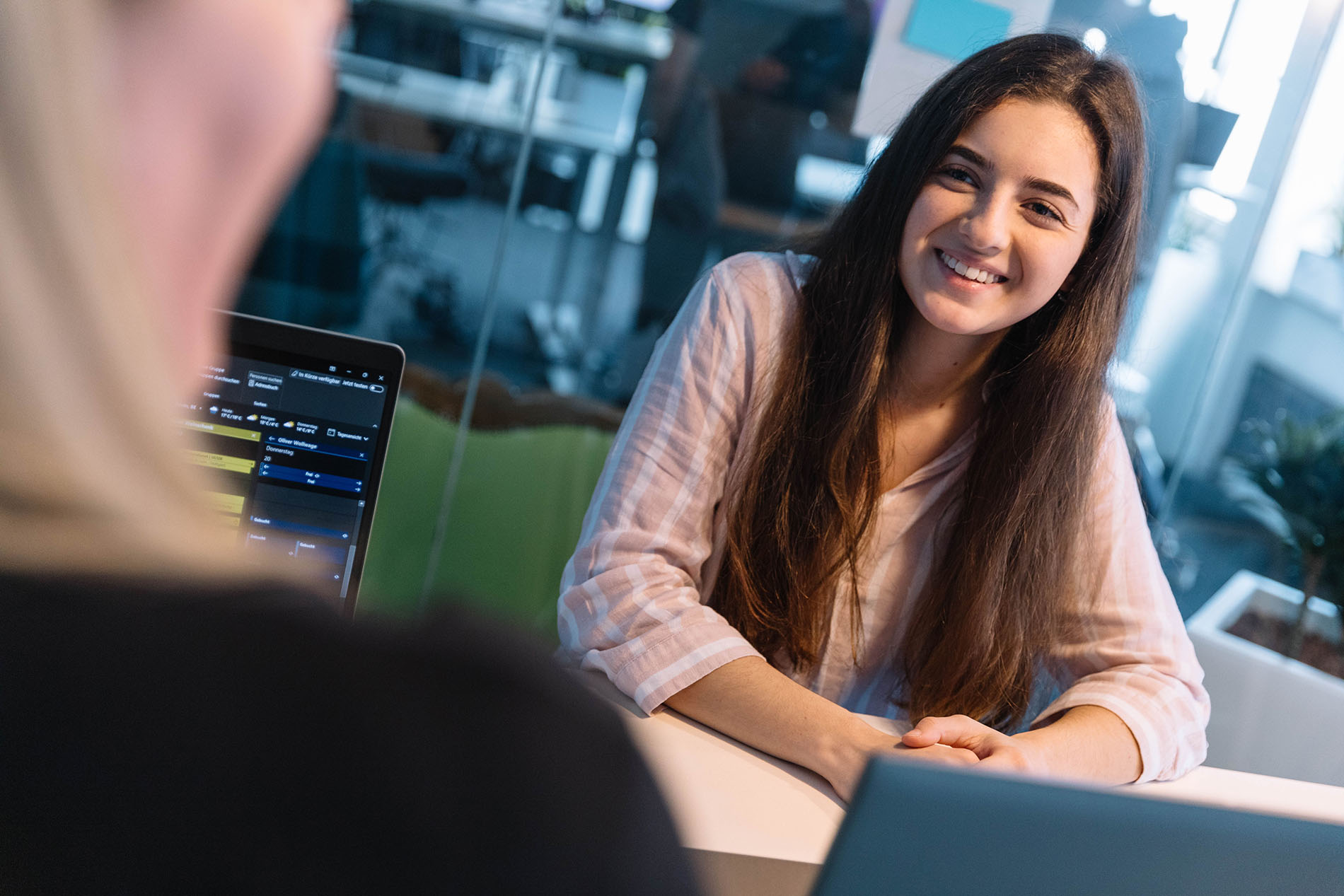 TYPO3-Agentur - Experten planen weiteres vorgehen Eine Frau mit langen dunklen Haaren sitzt an einem Tisch in einem modernen Büro und führt ein Gespräch, im Vordergrund steht ein Laptop.