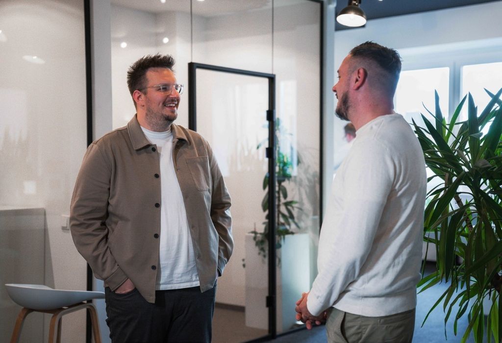 Zwei Mitarbeiter im Gespräch in einem modernen Büro mit Glaswänden und Pflanzen. Ein entspannter Austausch über ein laufendes Projekt.